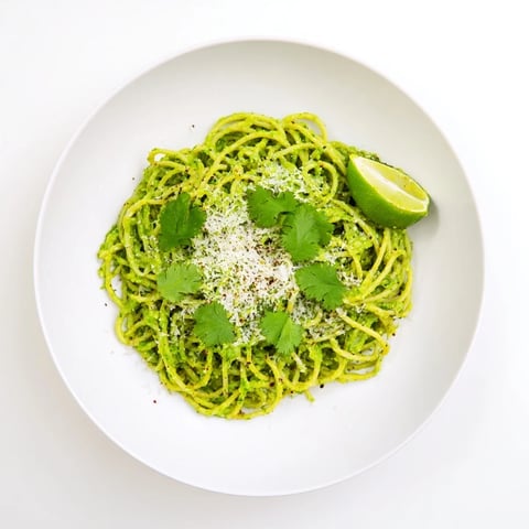 Creamy smashed avocado pasta twirls on a fork, garnished with fresh basil, lime wedges, and a sprinkle of chili flakes for a zesty vegetarian dinner.
