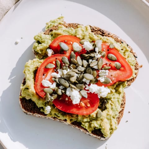 Golden avocado mash spread on crisp whole grain toast, topped with juicy tomato slices and crunchy pumpkin seeds.